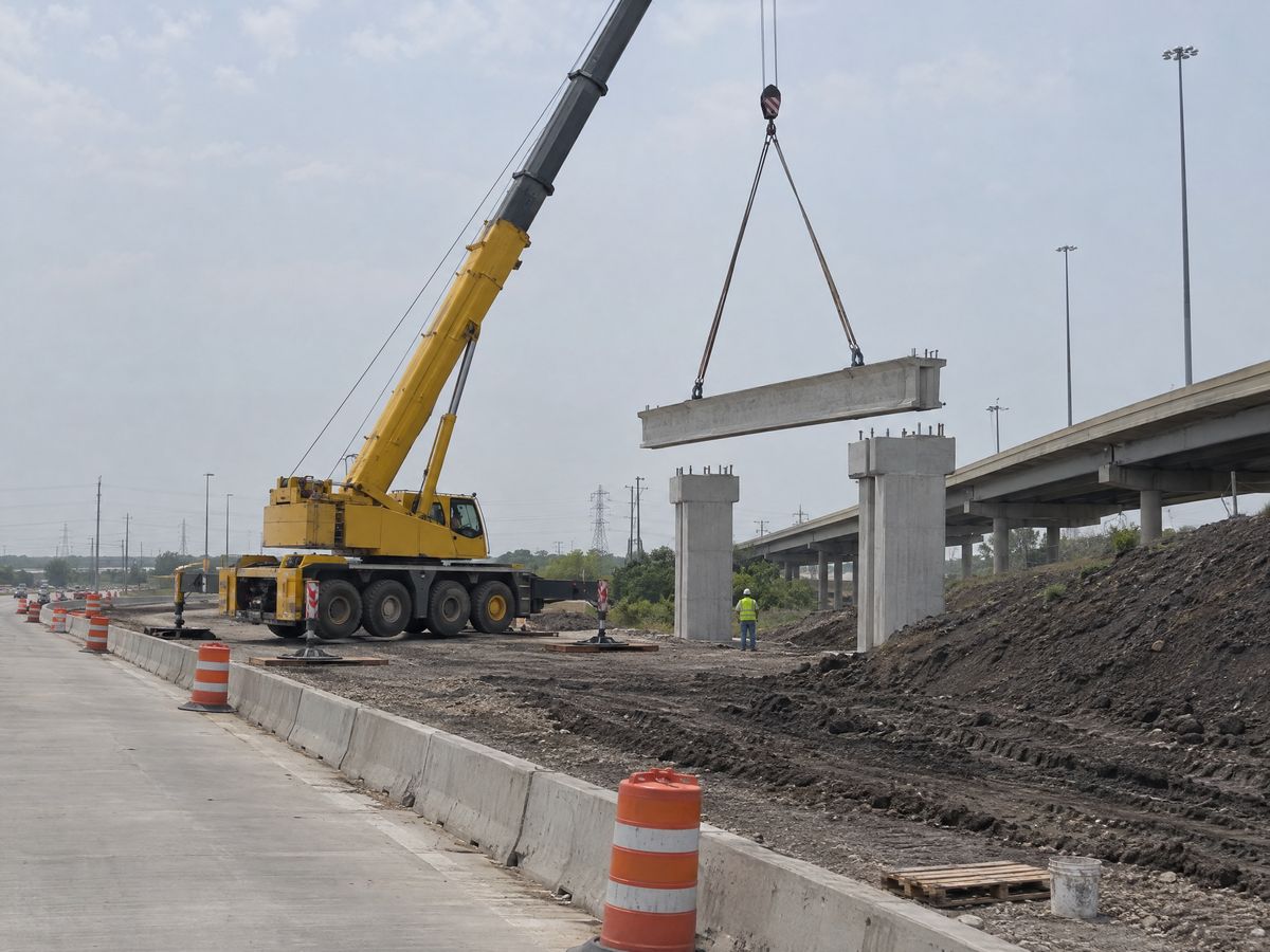 A yellow all-terrain crane on a highway frontage road swings a concrete girder toward new concrete bents beside an elevated highway.