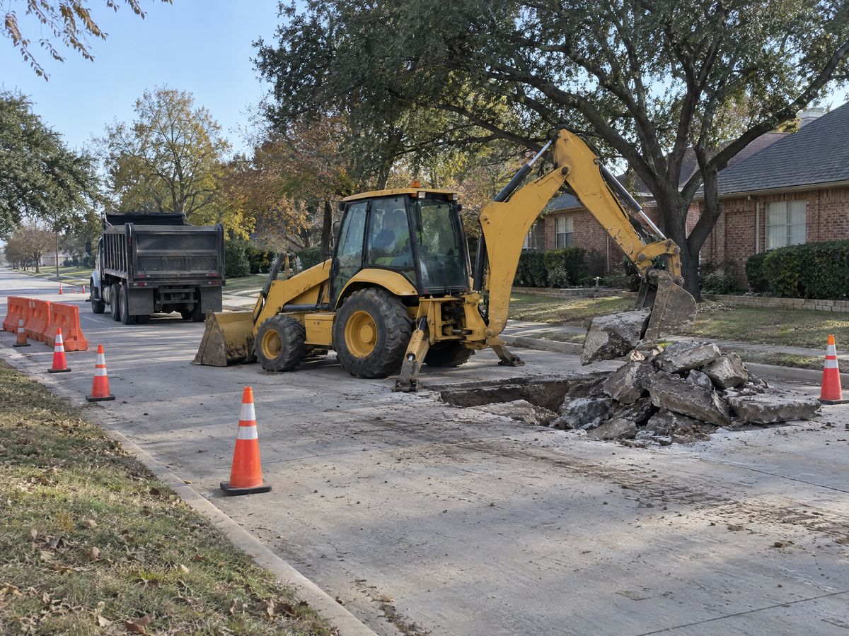 A yellow backhoe loader lifts broken concrete slabs from a cut in a residential street closed with cones, a dump truck waiting behind.