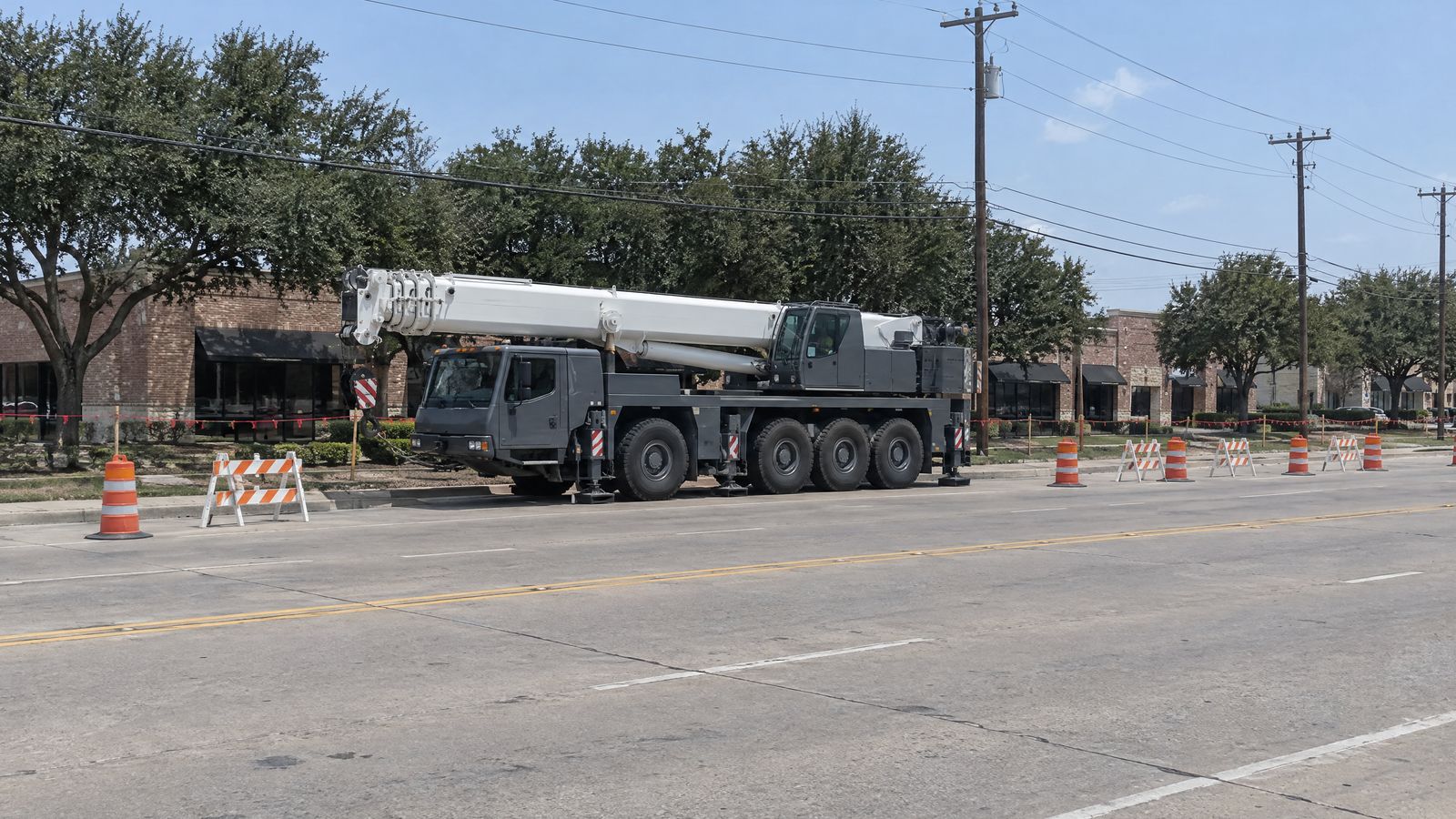 A grey crane with its boom stowed sits at the curb inside a lane closure of orange drums and barricades, under power lines on a street of brick shops.
