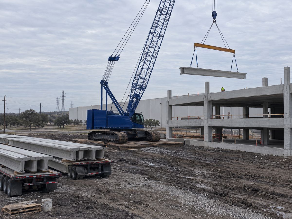 A blue lattice-boom crawler crane on timber mats lifts a precast double-tee beam on a spreader beam toward a parking garage under construction.