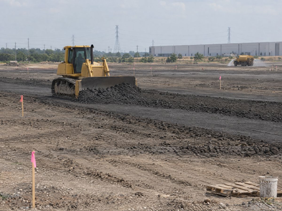 A yellow crawler dozer pushes dark clay across a large graded pad with pink-flagged stakes, a water truck and a warehouse in the distance.