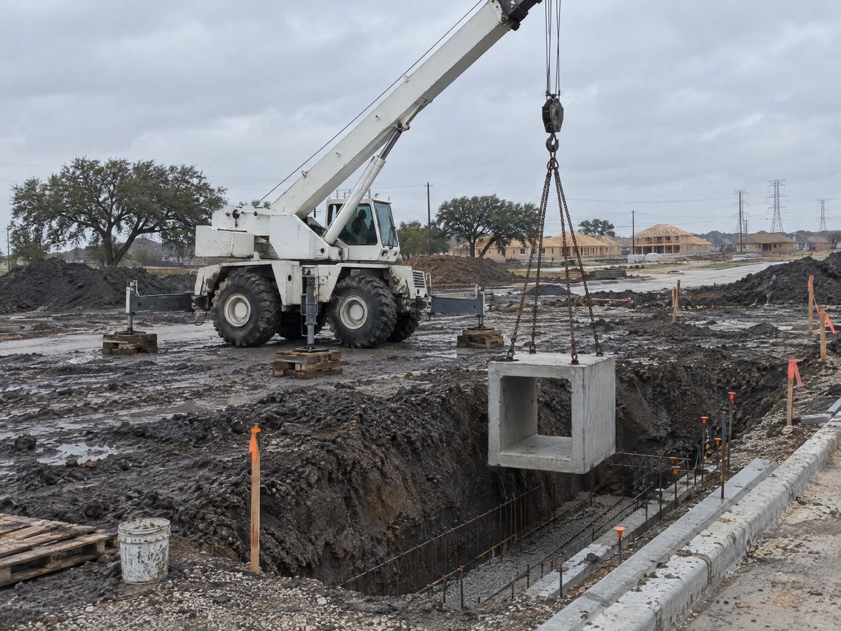 A white rough-terrain crane on timber cribbing lowers a precast concrete box section on chains into a muddy trench in a new subdivision.
