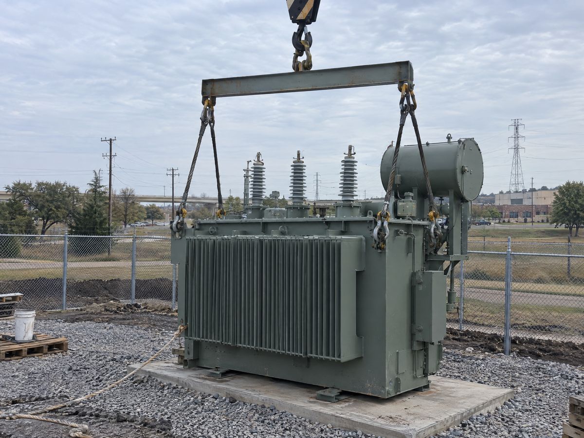 A steel spreader bar on a crane hook, with wire rope slings and shackles rigged to a large transformer on a concrete pad in a fenced yard.