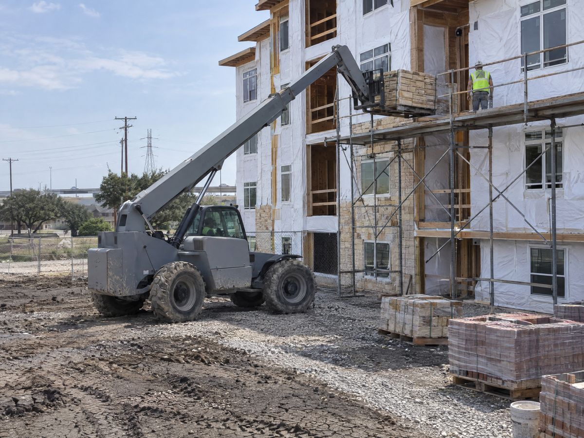 A grey telehandler sets a pallet of tan stone veneer on a scaffold at a wood-framed apartment building wrapped in white house wrap.