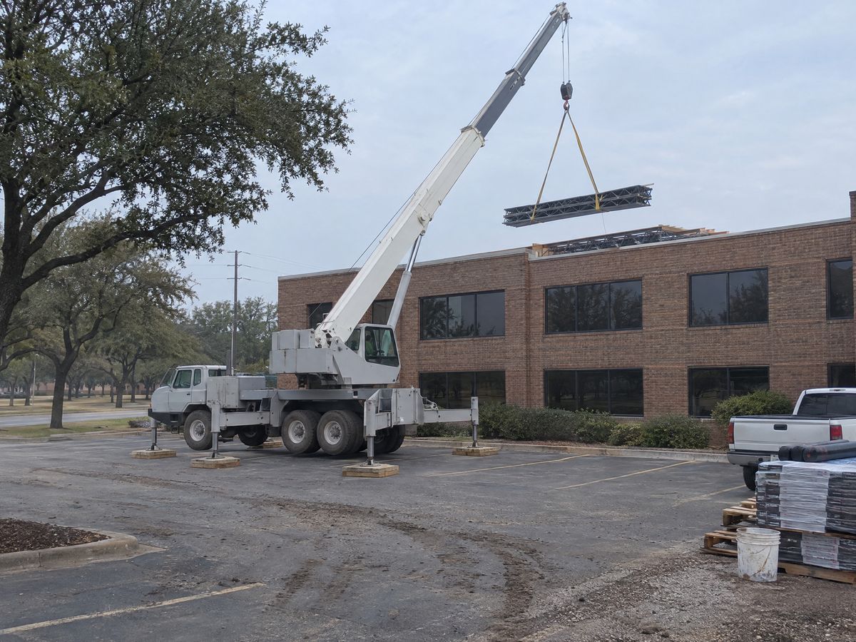 A light grey truck crane on timber outrigger pads lifts a bundle of steel joists onto the roof of a two-story brick office building.